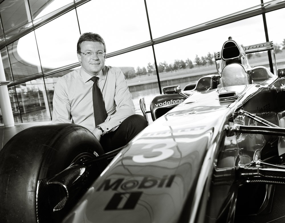 Black and white portrait photograph of Finance Director at Maclaren crouching over with F1 car at Mclaren HQ, Woking, Surrey