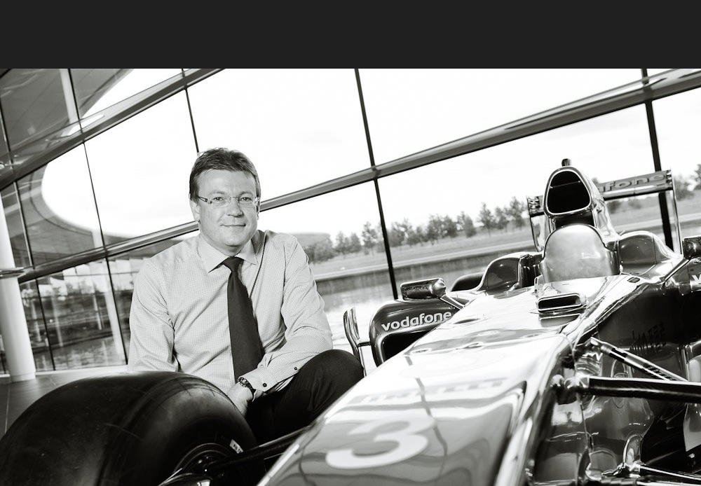 Black and white senior staff portrait - Mclaren Finanace Direcotr photo crouching by F1 car at Mclaren HQ